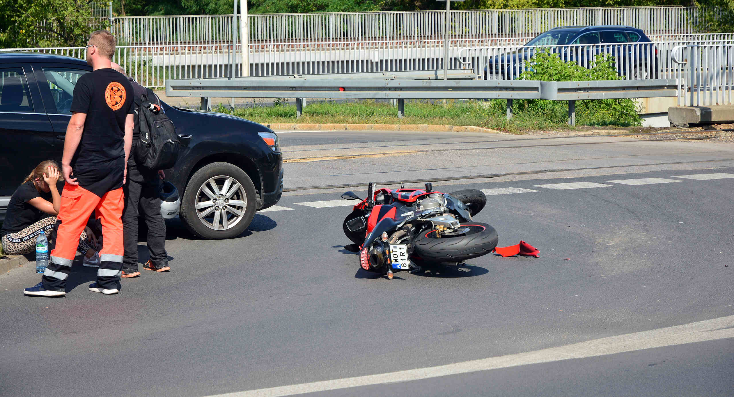 A red and black motorcycle lies on its side in a city intersection after a collision, with a car and bystanders nearby.